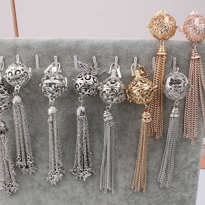 May include: Nine silver and gold cage-style pendants with tassel chains. The pendants are arranged in a row on a gray background.