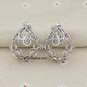 Könnte beinhalten: Zwei silberne Filigran-Käfig-Anhänger mit floralem Design. Die Käfige sind offen und können verwendet werden, um kleine Gegenstände wie Kristalle oder Charms aufzunehmen.