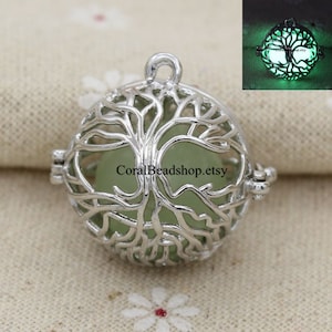 Puede incluir: Colgante de plata en forma de árbol de la vida con una piedra que brilla en la oscuridad en su interior. El colgante está abierto y la piedra es visible. El colgante está sobre un fondo de tela blanca.