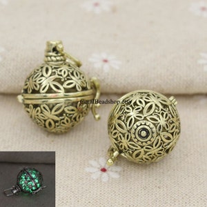 Peut inclure: Deux pendentifs en cage de métal doré avec des motifs floraux complexes. Les pendentifs sont ouverts et ont une petite boucle en haut pour les attacher à une chaîne. Les pendentifs sont sur un fond de tissu blanc. L'image du bas montre l'un des pendentifs qui brille en vert dans le noir.