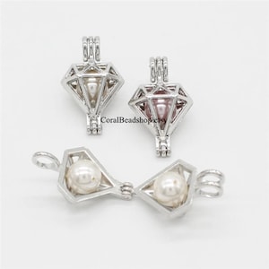 Puede incluir: Dos colgantes de plata en forma de diamante con una perla en su interior. Los colgantes están abiertos por un lado y se pueden usar como un medallón o un amuleto. Los colgantes están sobre un fondo blanco.