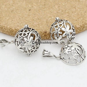 May include: Three silver-coloured, openwork pendants with floral designs. Each pendant is a sphere with a loop for hanging. The pendants are displayed on a light brown, textured surface.