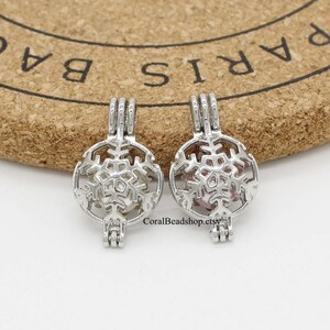 May include: Two silver-toned metal cage pendants featuring a snowflake design. Each pendant has a hinged top loop and a circular frame with a cutout snowflake pattern. A small, light-coloured spherical bead is visible inside each cage pendant.