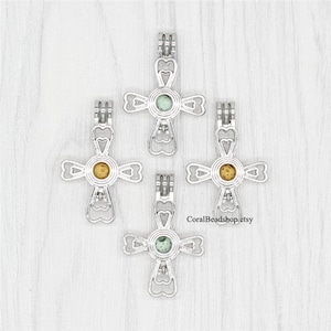 May include: Four silver cross-shaped charm pendants with a round opening in the centre. Each pendant has a different coloured pearl inside. The pearls are green, yellow, green, and green.