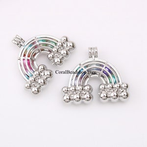 May include: Two silver-toned metal rainbow-shaped cage pendants featuring a multi-colored bead interior and cloud-like clusters at each end. The text CoralBeadshop.etsy is visible between the two pendants, which include a small bail at the top.