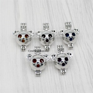 Puede incluir: Cinco jaulas para cuentas de metal plateado con forma de koala, cada una con una pequeña cuenta de color en su interior. Las jaulas tienen un diseño articulado con un lazo superior para colgar. Las cuentas son de color ámbar, rojo oscuro, azul oscuro, rojo brillante y morado.
