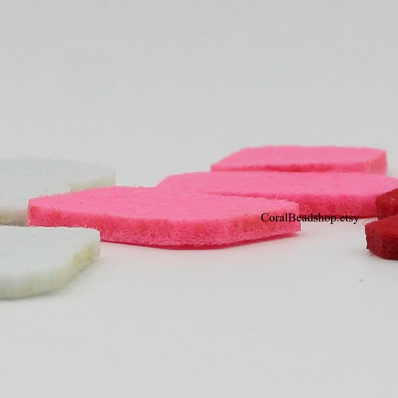 May include: A collection of small, flat, rectangular felt pieces in vibrant pink, white, and deep red colours. The felt shapes are arranged in a scattered formation, showcasing their soft, textured edges and uniform thickness. Text reads CoralBeadshop.etsy.