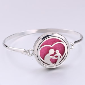 May include: Silver bangle bracelet with a round, silver locket featuring a cutout design of a mother and child inside a heart. The locket has a pink felt pad inside.