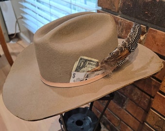 May include: A brown cowboy hat with a feather and a dollar bill tucked into the band. The hat is sitting on a black metal stand.