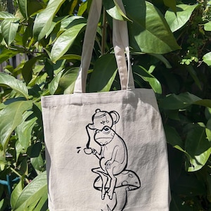 Puede incluir: Una bolsa de tela blanca con una ilustración en blanco y negro de una rana que lleva una tetera en la cabeza y sostiene una taza de té. La rana está sentada en un hongo.