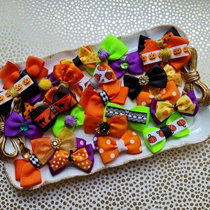 May include: A collection of Halloween-themed hair bows in various colors, including orange, purple, black, and green. Some bows feature pumpkin and bat designs, and are decorated with rhinestones and lace. The bows are arranged on a white decorative tray.