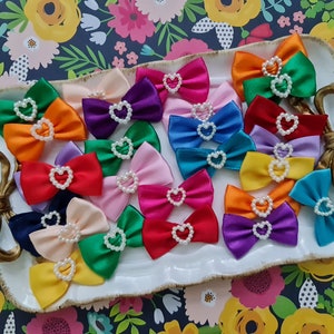 May include: A collection of 18 colorful satin hair bows with pearl accents in the shape of a heart. The bows are arranged on a white tray with a gold handle.