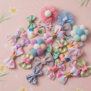 Op de afbeelding: Een verzameling kleurrijke haarstrikken in verschillende ontwerpen. De strikken hebben pastelkleuren, stippen, bloempatronen en prints met Pasen. Sommige strikken hebben glitteraccenten en pompomdetails. De strikken zijn gerangschikt op een roze ondergrond.
