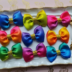 May include: Ten colourful satin ribbon bows with gold glitter centres. The bows are arranged in a rainbow pattern on a white tray.