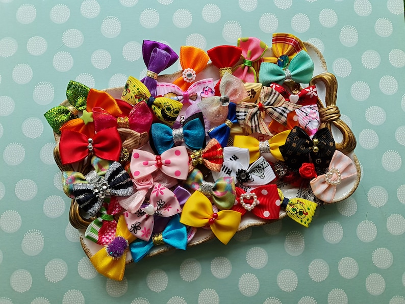 May include: A collection of colorful hair bows in various sizes and patterns, including polka dots, stripes, and floral designs. The bows are arranged on a decorative, gold-trimmed dish, set against a light blue background with white polka dots.
