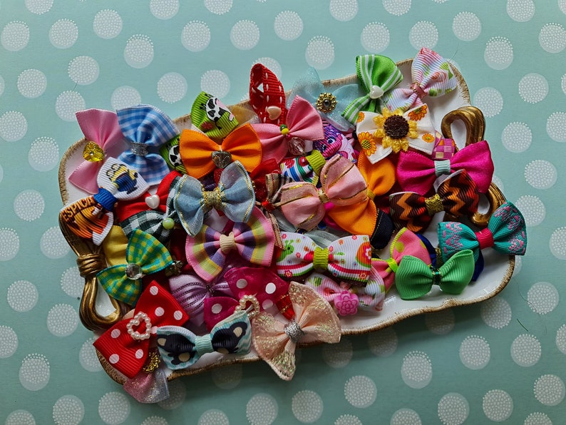 May include: A collection of colorful hair bows in various patterns and sizes, displayed on a decorative, off-white, heart-shaped dish with gold accents. The bows feature polka dots, stripes, and floral designs, with some embellished with rhinestones and pearls.