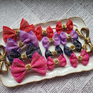 Puede incluir: Colección de lazos de tela coloridos en rojo, morado, azul marino y rosa, cada uno con un centro de purpurina dorada. Los lazos están dispuestos en una bandeja blanca adornada, para accesorios para el cabello.