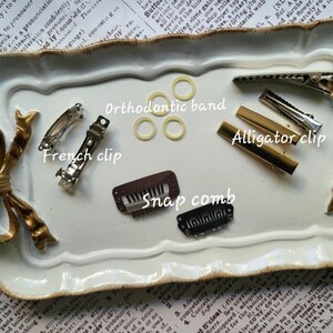 May include: A collection of hair accessories including French clips, alligator clips, snap combs, and orthodontic bands. The items are in silver, gold, and brown tones, arranged on a decorative white tray with gold accents.