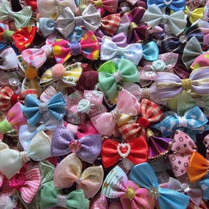May include: A collection of colorful hair bows in various sizes and designs. The bows feature a variety of colors, including pink, blue, green, and purple, with some adorned with rhinestones and other embellishments. The bows are made of different materials, such as satin and grosgrain.