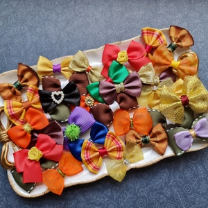 May include: A collection of colorful hair bows in various sizes and designs, including solid colors, patterns, and embellishments like pearls and rhinestones. The bows are arranged on a white, decorative tray with a gold handle, set against a blue floral background.