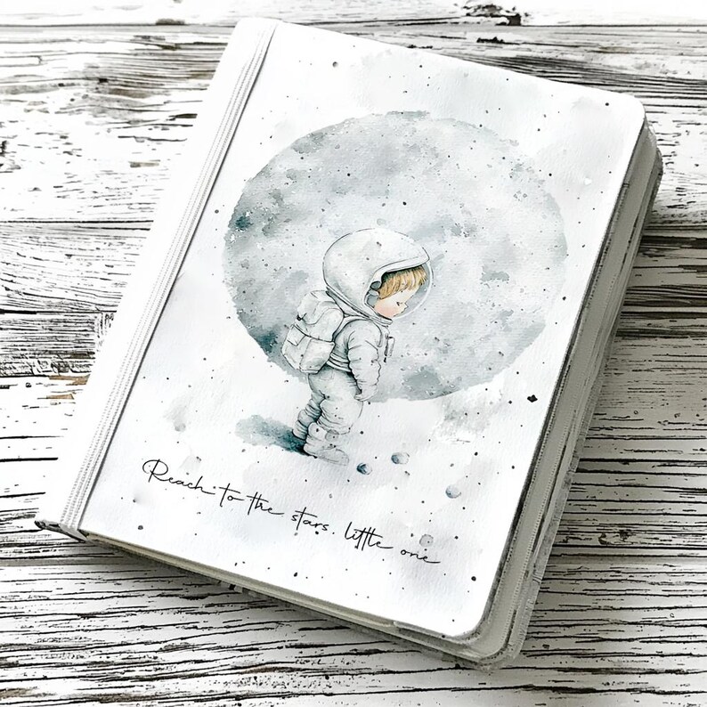 May include: White notebook with a watercolour illustration of an astronaut standing on the moon. The text "Reach to the stars, little one" is written on the cover.