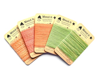 Könnte beinhalten: Fünf Spulen mit farbigem recyceltem Garn mit der Aufschrift "Mend it" und dem Text "Limited Edition Upcycled Yarn" sowie der Garnzusammensetzung unter jeder Spule. Die Spulen sind fächerförmig angeordnet.
