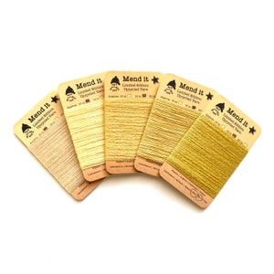 Pode incluir: Cinco rolos de fio reciclado amarelo e dourado etiquetados com "Mend it" com o texto "Limited Edition Upcycled Yarn" e o comprimento aproximado de cada rolo em metros.