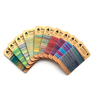 Op de afbeelding: Een verzameling kleurrijke garen strengen, elk met een unieke mix van tinten, gerangschikt in een waaier. Het garen is bevestigd aan bruine kartonnen labels met de tekst "Mend it" en "Limited Edition Upcycled Yarn".