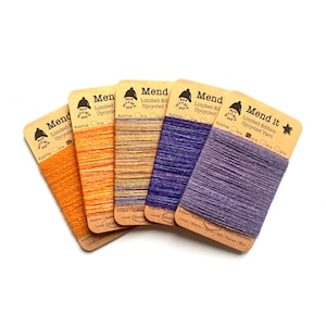 Op de afbeelding: Zes spoelen met kleurrijk garen met het label "Mend it Limited Edition Upcycled Yarn" met verschillende vezelmengsels, waaronder alpaca, merino, linnen, zijde en wol. De spoelen zijn ongeveer 10 meter lang.