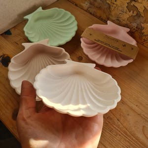 Può includere: Quattro piattini a forma di conchiglia in colori pastello: bianco, rosa, verde chiaro e un secondo piattino rosa con una fascia di carta marrone. I piattini sono in materiale liscio e poggiano su una superficie di legno.
