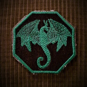 May include: A green embroidered dragon patch on a brown fabric background. The dragon is in a flying pose with its wings spread out.
