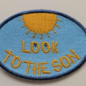 May include: An oval-shaped blue embroidered patch with a yellow sun and the text "LOOK TO THE SUN" in yellow thread.