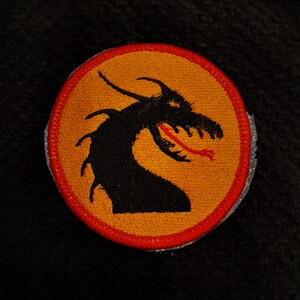 May include: A round orange patch with a red border, featuring a black silhouette of a dragon's head with a red tongue.
