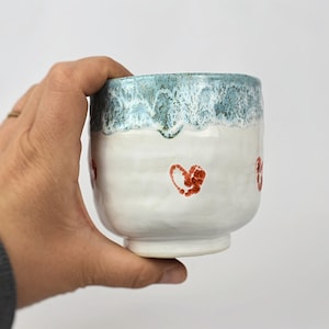 May include: A handmade ceramic cup with a white base and a turquoise rim. The cup features red heart designs. The cup is held in a hand, showcasing its size and shape. The cup is likely used for drinking beverages.