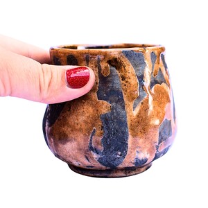 Könnte beinhalten: Eine kleine, handgefertigte Keramiktasse mit einem einzigartigen, organischen Design. Die Tasse weist eine Mischung aus braunen, schwarzen und beigen Farben auf, die einen Marmoreffekt erzeugt. Die Tasse wird von einer Hand mit rotem Nagellack gehalten.