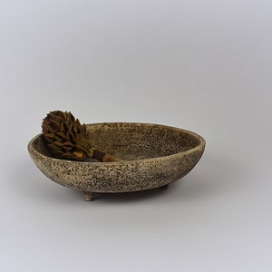 Peut inclure: Un petit bol en céramique de forme ovale avec un glaçage texturé brun et gris moucheté. Le bol a trois petits pieds et repose sur une surface blanche. Un bourgeon de fleur de magnolia séché repose à l'intérieur du bol.