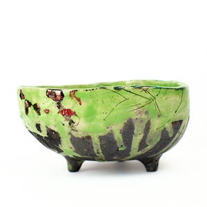 May include: A handcrafted ceramic bowl with a bright green glaze and dark brown detailing. The bowl has an unusual, uneven form and rests on three small feet. The inside is a lighter green, and the outside has a textured, artistic pattern.