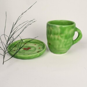 Könnte beinhalten: Ein grünes Keramik-Teetassen- und Untertassen-Set mit einem gesprenkelten Überzug und roten Akzenten. Die Untertasse hat einen leicht gewellten Rand.