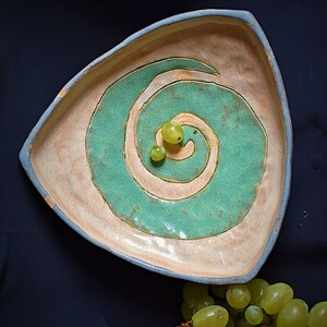 Puede incluir: Un plato de servir triangular de cerámica con un glaseado marrón claro y un diseño de espiral verde. El plato está decorado con un borde azul claro. Dos uvas verdes están en el centro del plato.