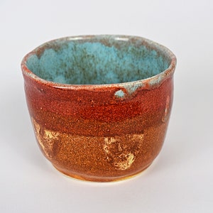 May include: A small, round ceramic bowl with a teal glaze on the inside and a brown, red, and yellow glaze on the outside. The bowl has a rustic, handmade look.
