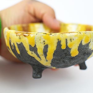 Puede incluir: Un pequeño cuenco de cerámica hecho a mano con un diseño único. El cuenco presenta una base gris oscuro con un esmalte amarillo vibrante que gotea por los lados. Tiene tres pequeños pies para mayor estabilidad. Ideal para servir pequeñas porciones.