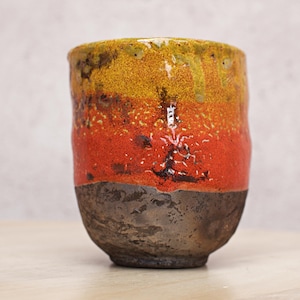 Peut inclure: Un gobelet en céramique fait à la main avec une surface texturée. La tasse présente un dégradé de couleurs, passant d'une base sombre et terreuse à un milieu rouge vif, et surmontée de teintes jaunes et brunes. Le design est rustique et unique.