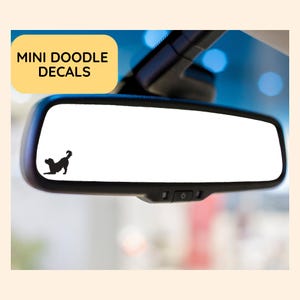 May include: A black silhouette of a dog decal is on a car rearview mirror. The decal is on the left side of the mirror. The text "MINI DOODLE DECALS" is in a yellow box in the upper left corner.
