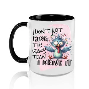 May include: White ceramic mug with a black handle and rim. The mug features a cartoon bird with the text "I DON'T JUST RIDE THE CRAZY TRAIN I DRIVE IT". The bird is blue with a red tongue and is set against a pink background.