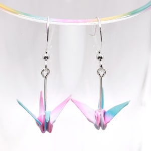 May include: Origami crane earrings with silver-coloured hooks and beads. The paper cranes are in shades of pink, blue, and white. The earrings are displayed inside a clear glass. The earrings are a delicate accessory.
