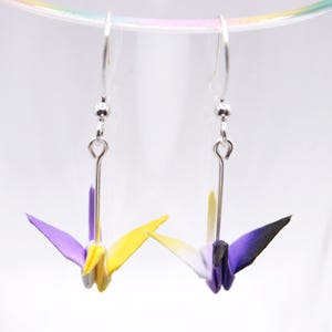 May include: Origami crane earrings, each featuring a paper crane in shades of purple, yellow, and white. The earrings hang from silver-coloured hooks. The background is a soft, out-of-focus white.