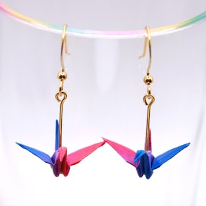 May include: Gold-coloured dangle earrings with origami crane pendants. The paper cranes are blue and pink, suspended from gold-coloured hooks. The earrings are set against a white background.