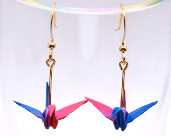 May include: A pair of gold-colored dangle earrings featuring origami crane pendants. The paper cranes are blue and pink, with gold-colored hooks. The earrings are displayed against a white background.