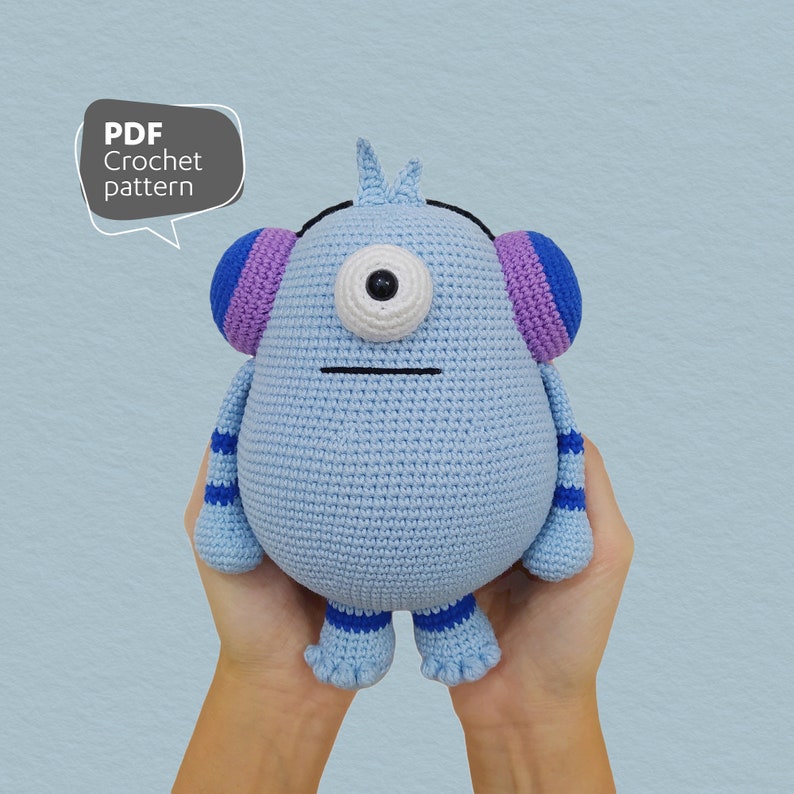 May include: A light blue crocheted monster with a single large eye, purple and blue earmuffs, and blue and white striped legs. The monster is being held by two hands.