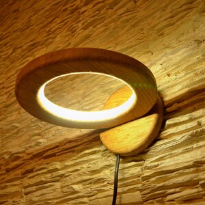 Könnte beinhalten: Eine Wandleuchte aus Holz mit einem runden Leuchtkörper. Die Leuchte ist aus Naturholz gefertigt und hat ein warmes, einladendes Aussehen. Der Leuchtkörper ist ein Ring aus LED-Leuchten, die ein weiches, weißes Licht abgeben.